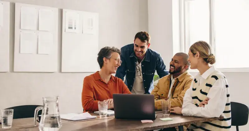 Four colleagues smiling and collaborating around a laptop at a shared office table
