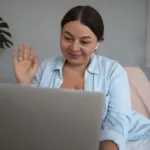 Guía práctica para integrar desarrolladores remotos a tu equipo con éxito Desarrolladora trabajando en su computadora y saludando durante su primera semana de onboarding remoto